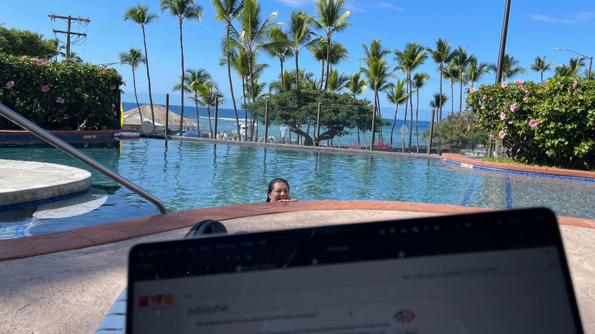 woman in pool visible over laptop screen; palm trees and ocean is in the background