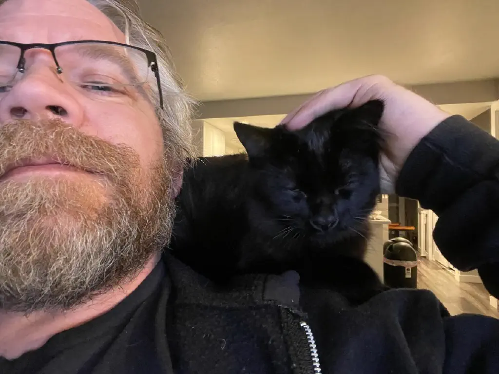 Extreme nose hair selfie of beardy dude with extremely content black kitty getting skritches on his shoulder