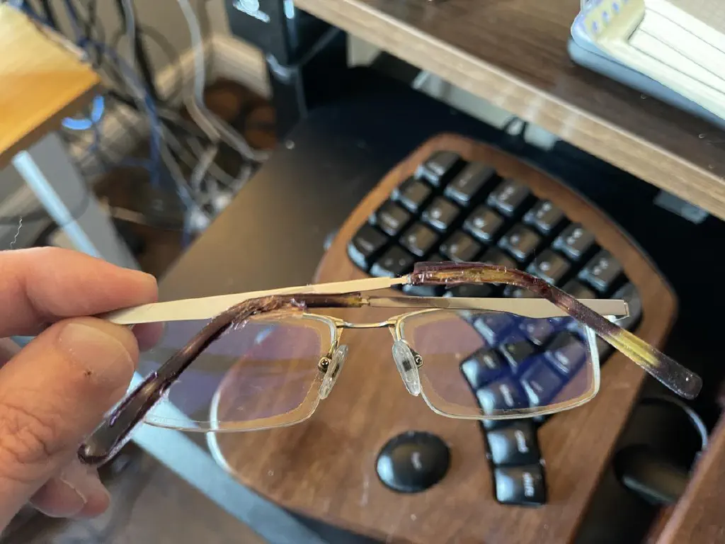 Hand holding a pair of metal frame glasses, with plastic earpieces that have partially melted.