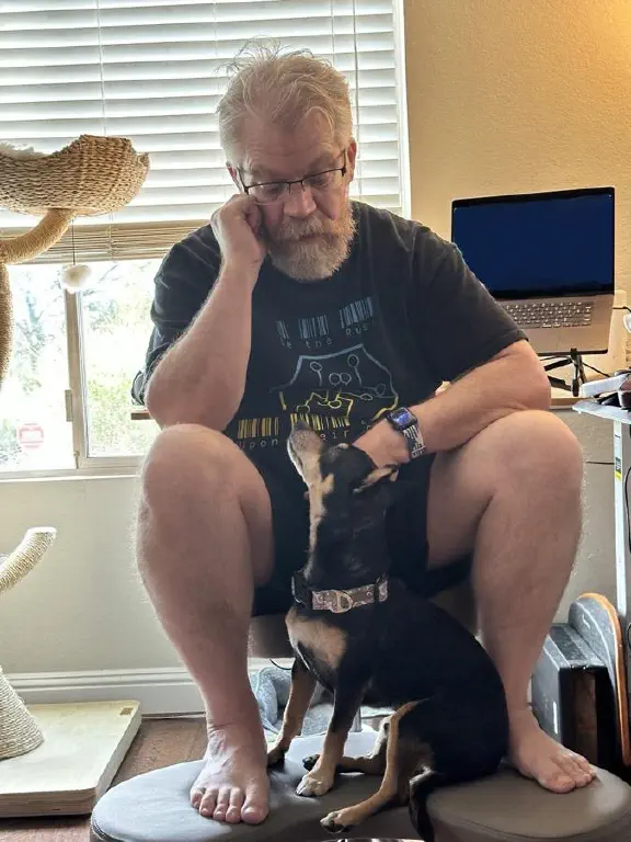 Black chihuahua mix dog sits on a footrest connected to an office chair, between the legs of a beardy nerd dude; they’re both looking at each other