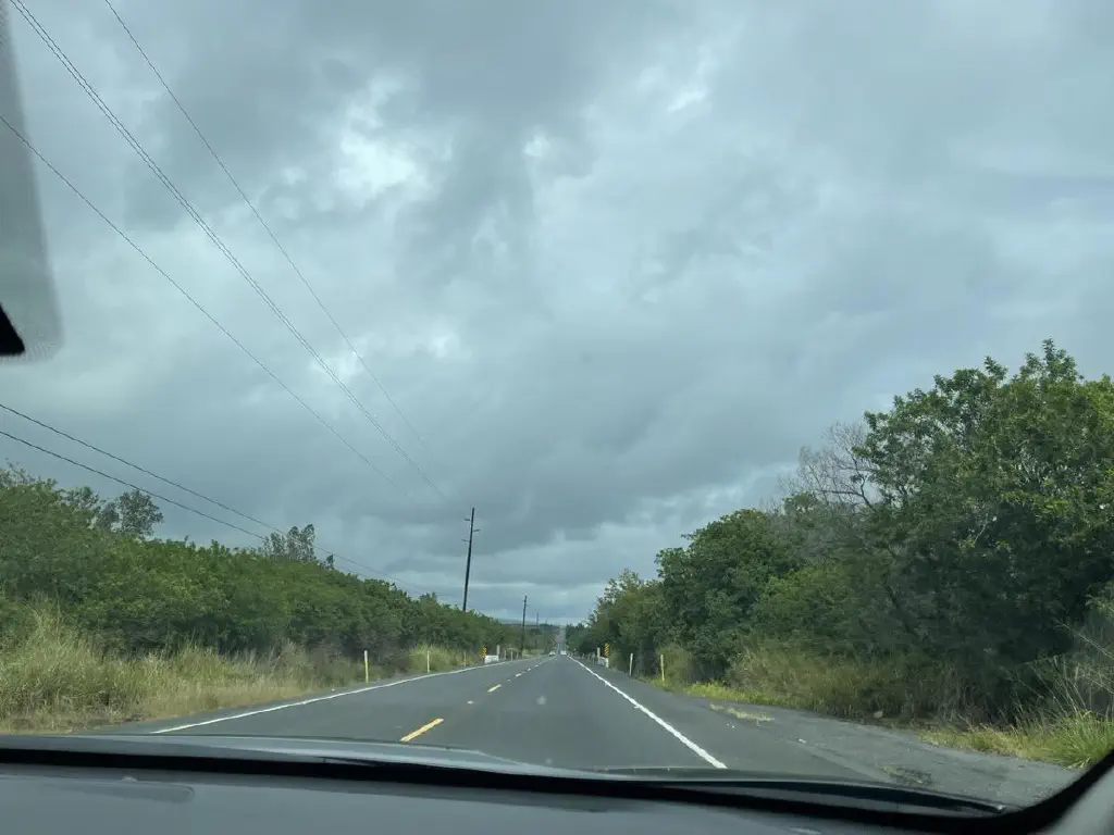 Photo from car of two lane road with tropical foliage on the sides and thick clouds overhead