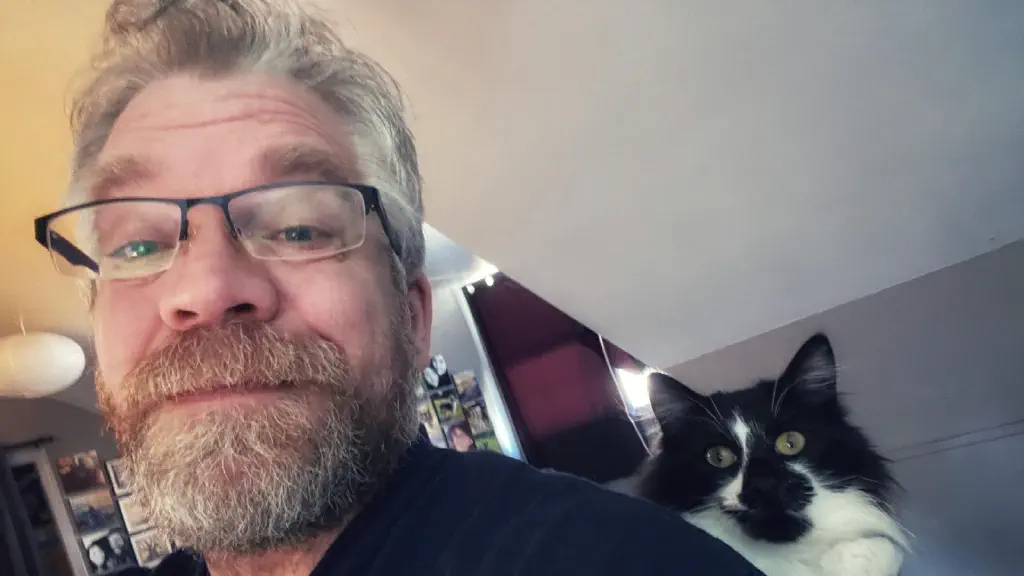 Selfie of beardy dude wearing glasses; sitting comfortably behind his shoulder is Jack, a black and white cat