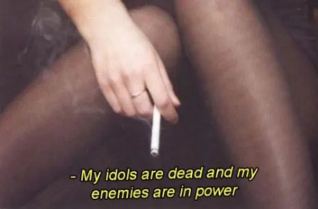 arm resting on stocking legs, hand lazily holding a lit cigarette. Text reads as if movie caption: “- My idols are dead and my enemies are in power”