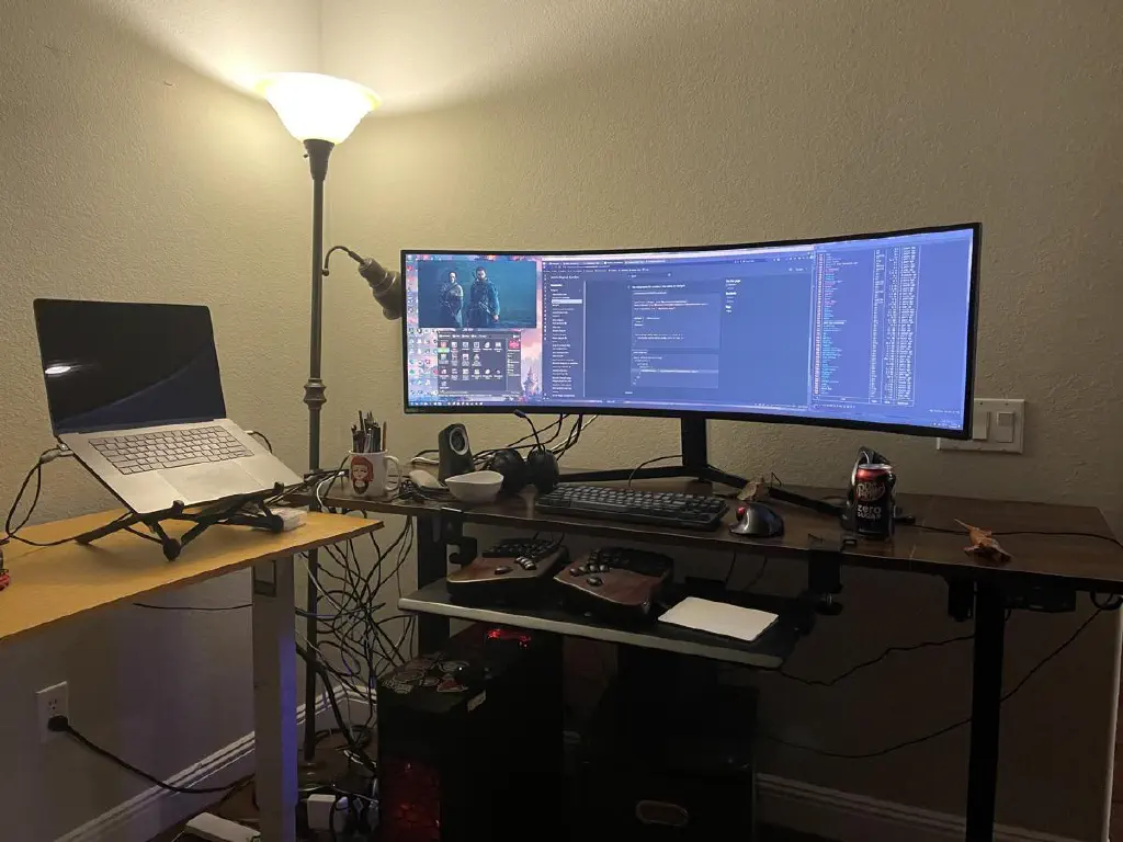 Two adjustable desks placed kitty corner to each other. One has an ultrawide monitor and two keyboards under it. The other has a MacBook Pro open on a laptop stand.
