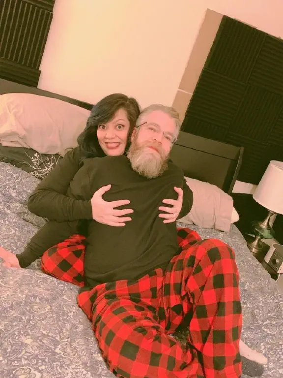 Beardy dude and dark haired woman lounging in pajamas, her grinning silly with hands on his chest
