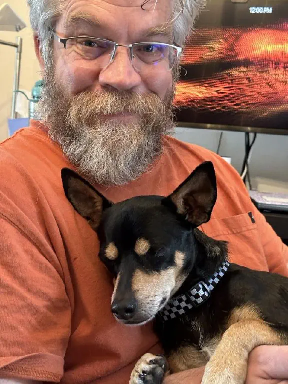 An extremely contented small dog rests in the arms of a smiling beardy dude