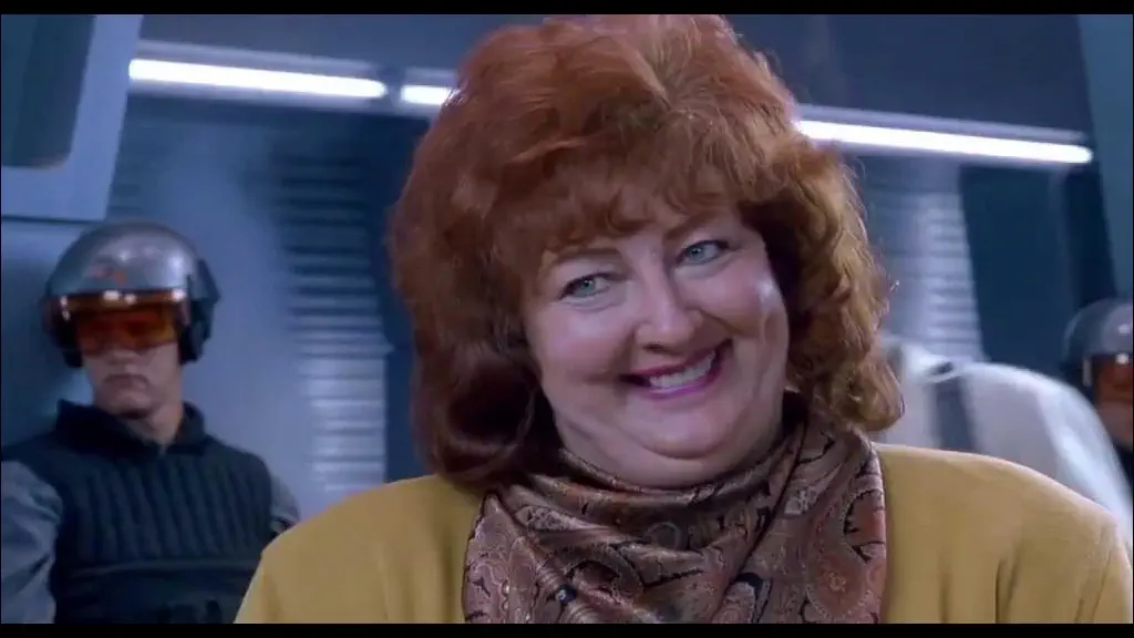 middle-aged woman with red hair offering a congenial smile in a sci-fi setting; from “Total Recall”