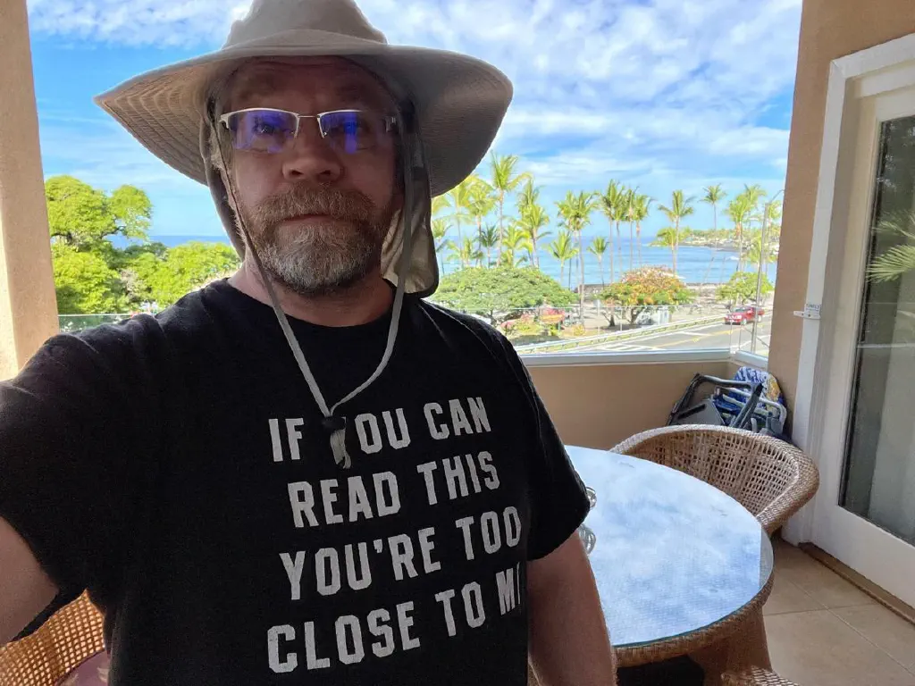 Background: sunny ocean beach with palm trees: foreground: bearded dude in glasses, floppy hat, and t-shirt that reads “if you can read this you’re too close to me”