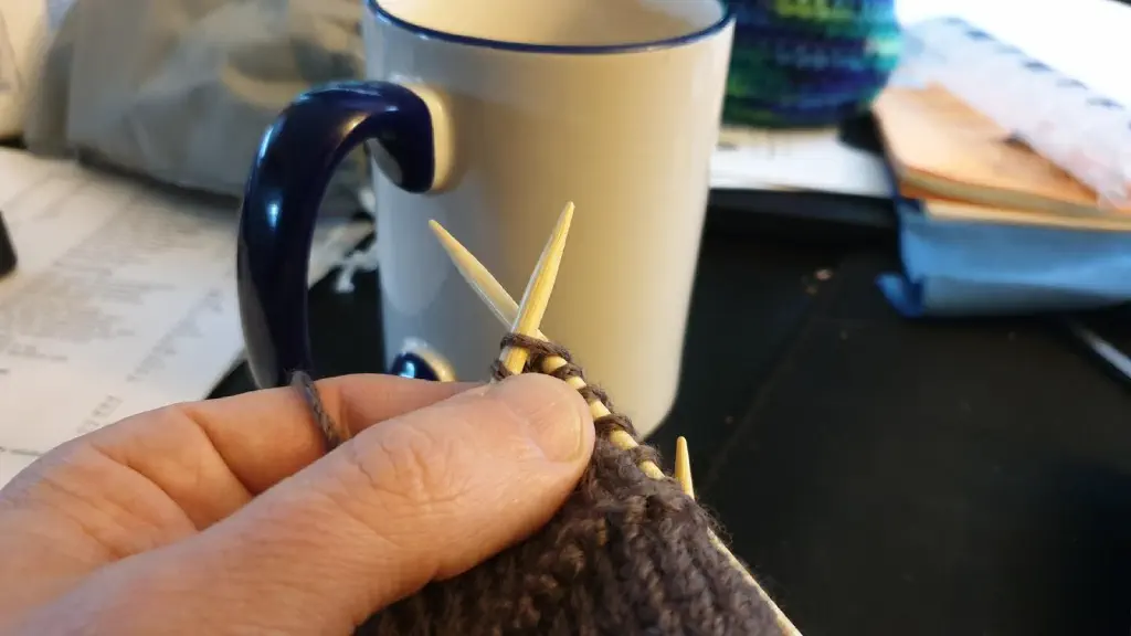 Foreground: hand holding knitted project mid-stitch; background: coffee