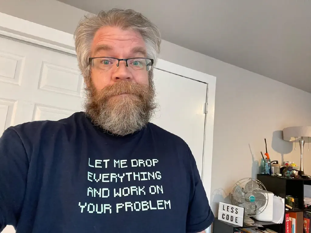 Bleary-eyed morning selfie of beardy dude in t shirt that reads “let me drop everything and work on your problem”