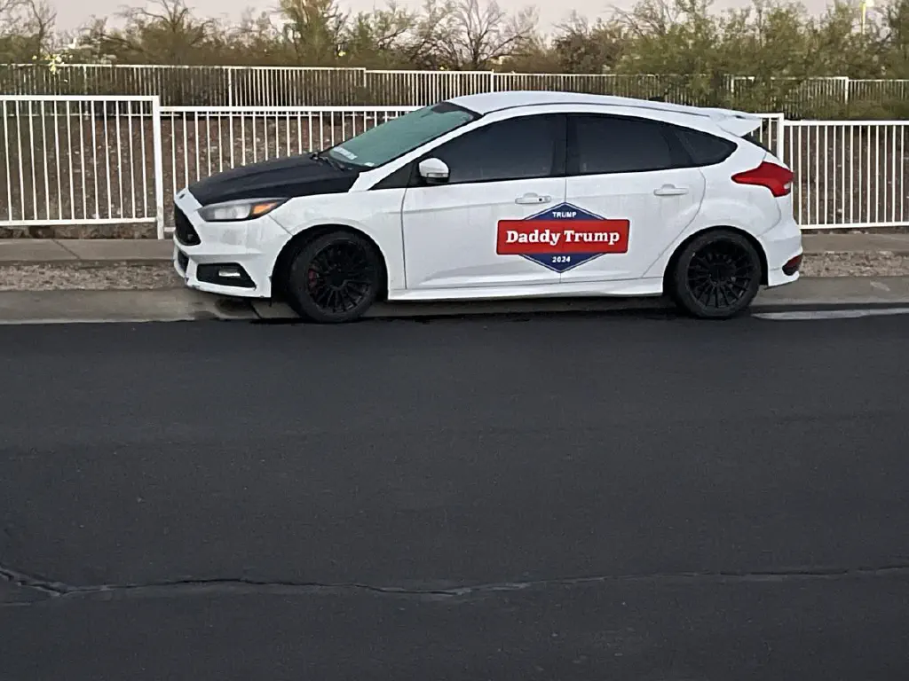 White car with red and blue decal on the side; decal text reads “Daddy Trump” and “Trump 2024”