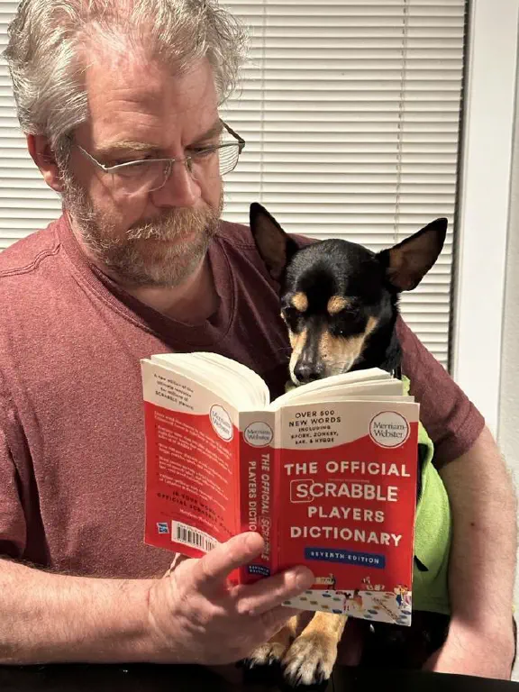 scruffy middle aged nerd in glasses shows an open copy of &ldquo;The Official Scrabble Players Dictionary&rdquo; to a small black chihuahua mix dog in his lap; the dog graciously pretends to be interested