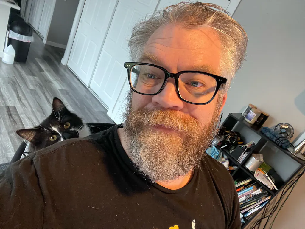 Selfie of beardy dude with glasses, black and white cat calmly poking head up from his resting place wedged behind beardy dude in office chair