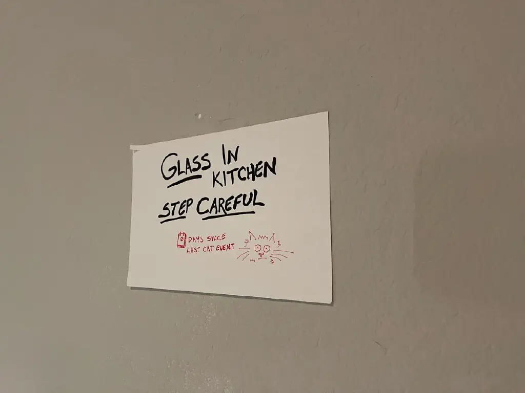 Sketchbook paper taped to wall: large black text reads “GLASS IN KITCHEN STEP CAREFUL” in black, with “0 days since last cat event” and quick doodle of a startled-looking cat in red