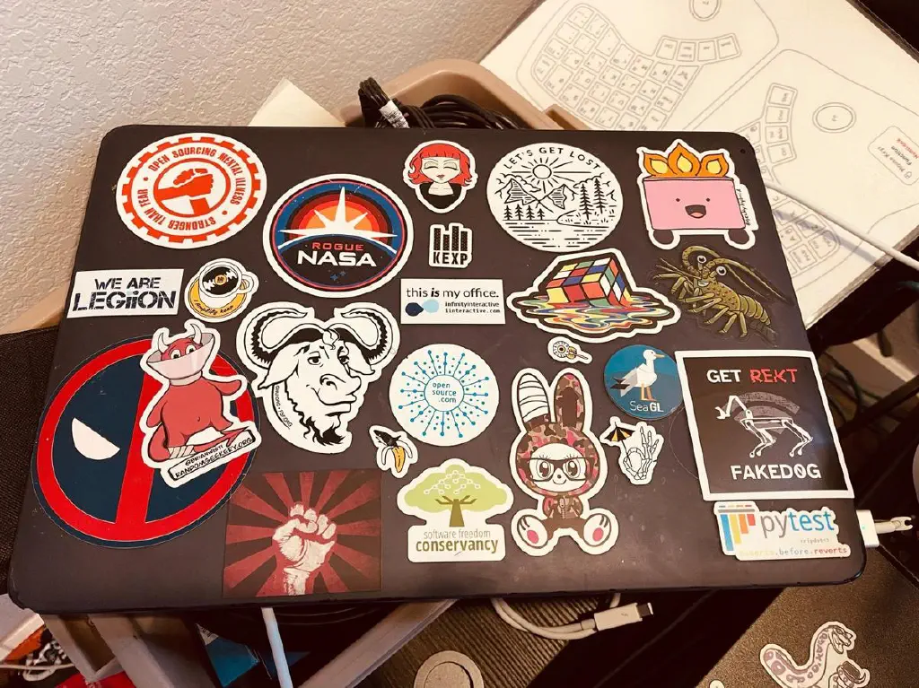 Laptop covered with stickers. New one has image of Boston Dynamics robot dog with “GET REKT FAKED0G” in big hostile letters
