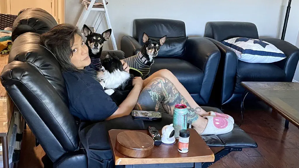 Woman sits on a couch, with a cat in her. Lap and two very alert black chihuahua mix dogs right next to her on the couch