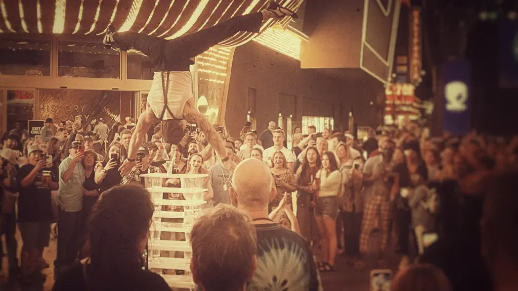 A street performer doing a headstand with arms and legs out, head resting on almost 2 meters of stacked plastic racks; a large crowd watches