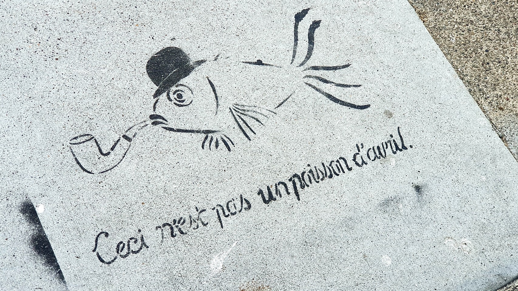 Stencil graffiti on sidewalk of fish with pipe and wearing bowler hat. Caption underneath: “Ceci n’est pas un poisson d’avril.”