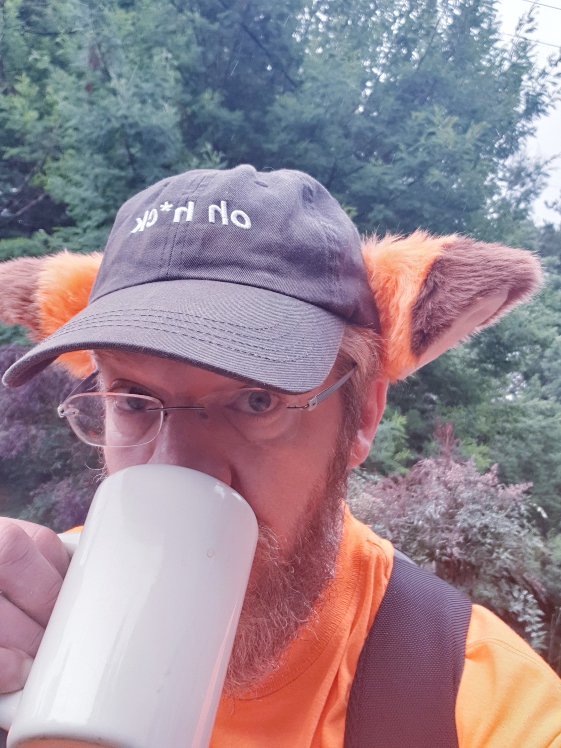 selfie with coffee: cat-fox ears pinned to a worn black baseball cap with “oh h*ck” embroidered in white