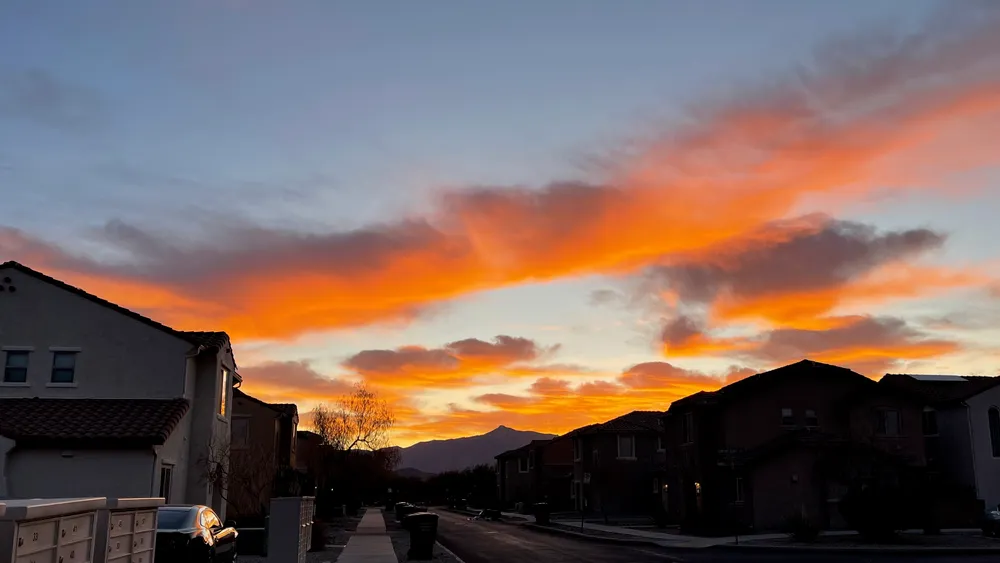 Dawn sky over suburban housing development in the desert, with clouds tinted orange and violet by a sun still hidden behind mountains