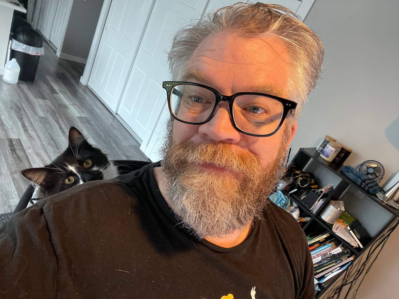 Selfie of beardy dude with glasses, black and white cat calmly poking head up from his resting place wedged behind beardy dude in office chair