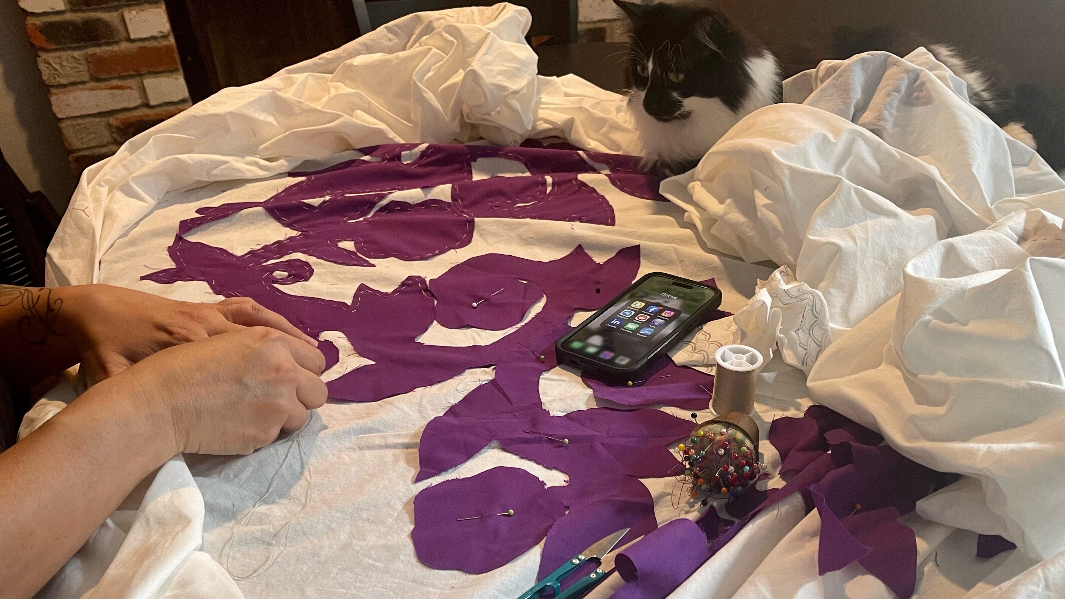 A cat sits on the edge of a quilt, watching it get stitched 